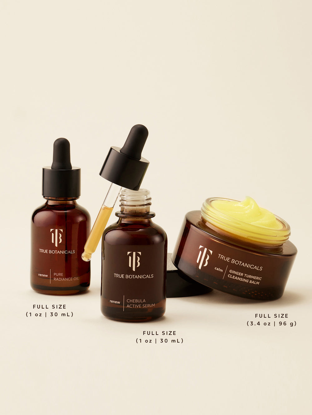 Three full-size True Botanicals skincare essentials — Pure Radiance Oil, Chebula Active Serum, and Ginger Turmeric Cleansing Balm — shown on a soft, neutral background. The rich golden balm is visibly textured in its open jar, while the oil and serum display their nourishing, plant-powered formulas in amber glass bottles with droppers. Sizes include 1 oz (30 mL) for both the oil and serum, and 3.4 oz (96 g) for the cleansing balm.
