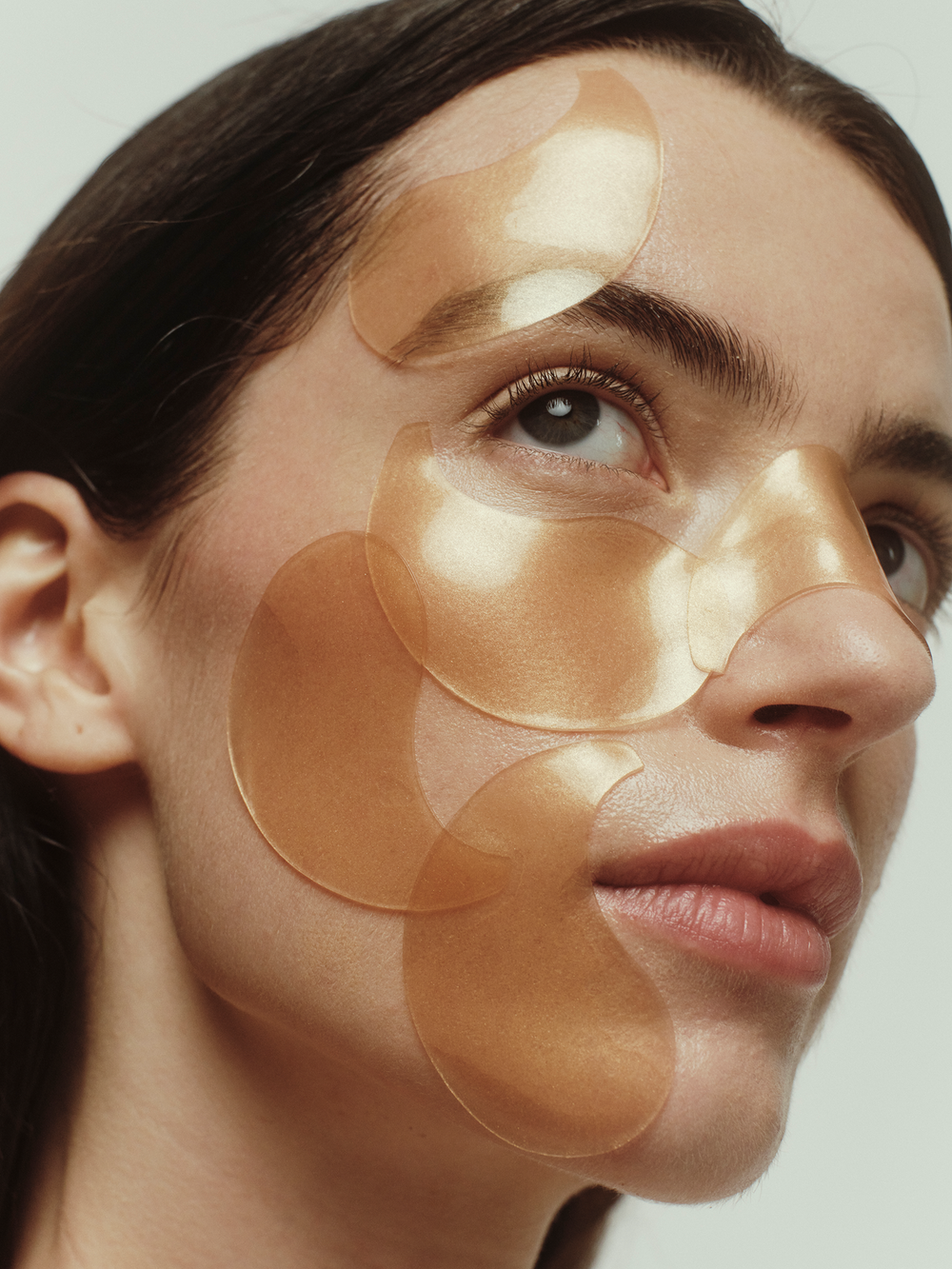 Chebula Multi-Corrective Eye Patches by True Botanicals shown applied to a model's face, with multiple translucent amber-toned gel patches layered across the eye area, cheekbones, and forehead against a soft neutral background.