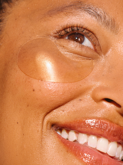 Chebula Multi-Corrective Eye Patches by True Botanicals shown applied under the eye of a smiling model with medium-deep skin tone, with a single translucent oval gel patch catching warm golden light against glowing skin.