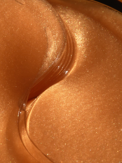 Close-up texture shot of True Botanicals Chebula Multi-Corrective Eye Patches gel formula, showing a rich copper-gold gel with iridescent shimmer and a glossy, folded edge detail.