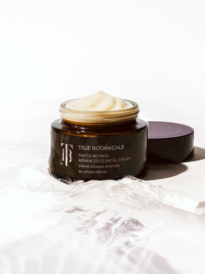 Open jar of True Botanicals Phyto-Retinol Advanced Clinical Cream showing a smooth, whipped texture, with the lid beside it and a glossy, water-like surface in the foreground.