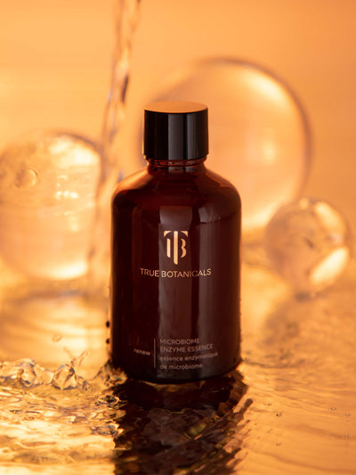 True Botanicals Microbiome Enzyme Essence in an amber glass bottle, surrounded by golden liquid and soft, glowing orbs, highlighting a fluid, radiant texture.